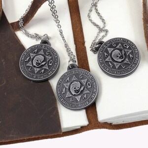 May include: Three silver-tone pendant necklaces with circular pendants featuring an engraved design and text. The pendants hang on delicate chains. The necklaces rest on a white surface next to a brown leather journal.