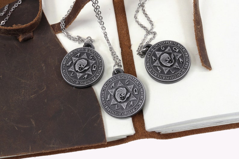 May include: Three silver-tone pendant necklaces with circular pendants featuring an engraved design and text. The pendants hang on delicate chains. The necklaces rest on a white surface next to a brown leather journal.