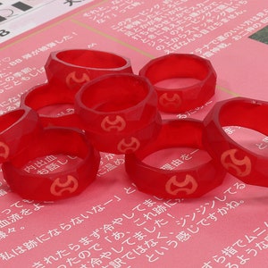 Op de afbeelding: Een verzameling gefacetteerde, doorschijnende rode ringen met een gouden embleem. De ringen liggen op een roze oppervlak met Japanse tekst. De ringen lijken van hard plastic of hars te zijn.