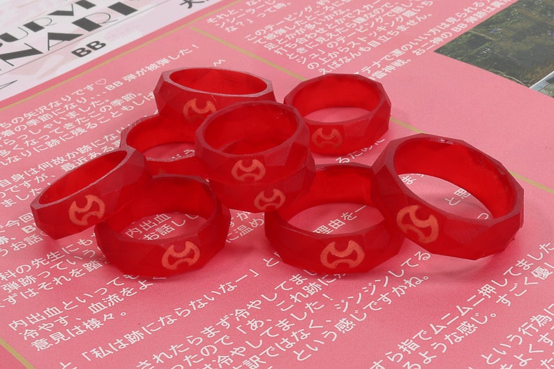 Op de afbeelding: Een verzameling gefacetteerde, doorschijnende rode ringen met een gouden embleem. De ringen liggen op een roze oppervlak met Japanse tekst. De ringen lijken van hard plastic of hars te zijn.
