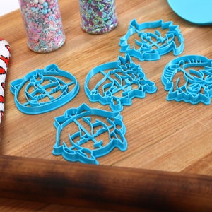 Pu&ograve; includere: Cinque tagliabiscotti in plastica blu a forma di facce di cartoni animati su una superficie di legno. I tagliabiscotti sono disposti a semicerchio con un mattarello e una spatola sullo sfondo.