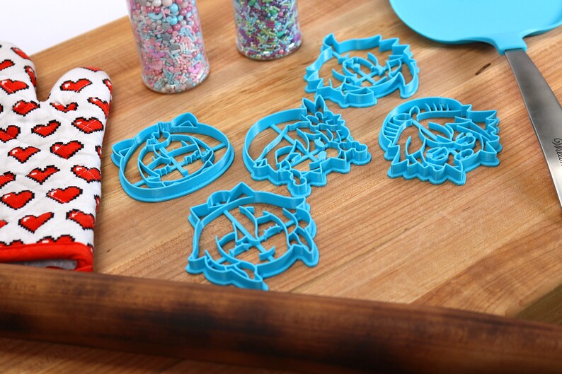 Pu&ograve; includere: Cinque tagliabiscotti in plastica blu a forma di facce di cartoni animati su una superficie di legno. I tagliabiscotti sono disposti a semicerchio con un mattarello e una spatola sullo sfondo.