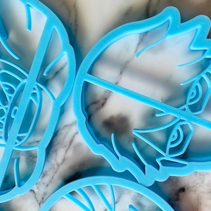 May include: Three blue plastic cookie cutters, one shaped like a face with a closed eye, one shaped like a face with an open eye, and one shaped like a swirl.