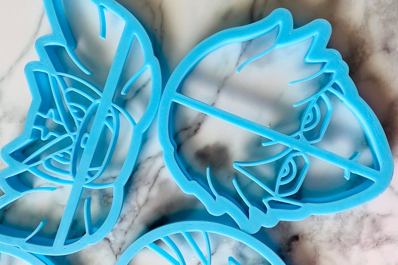 May include: Three blue plastic cookie cutters, one shaped like a face with a closed eye, one shaped like a face with an open eye, and one shaped like a swirl.