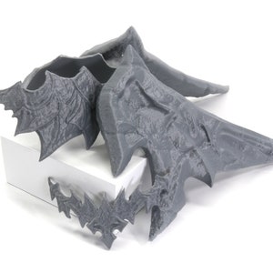 May include: Four grey 3D printed wings with intricate details. The wings are arranged in a pile on a white surface.