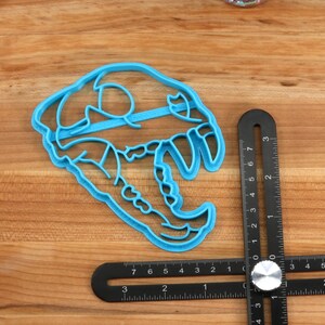May include: A blue plastic cookie cutter shaped like a dinosaur skull. The cutter is 3 inches wide and is sitting on a wooden surface next to a ruler.
