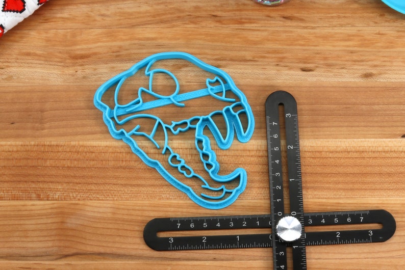 May include: A blue plastic cookie cutter shaped like a dinosaur skull. The cutter is 3 inches wide and is sitting on a wooden surface next to a ruler.