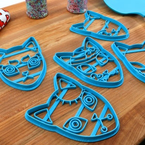 K&ouml;nnte beinhalten: Vier blaue, plastikene Ausstecher in Katzenform auf einer Holzoberfl&auml;che. Jeder Ausstecher hat ein anderes Design mit filigranen Details.