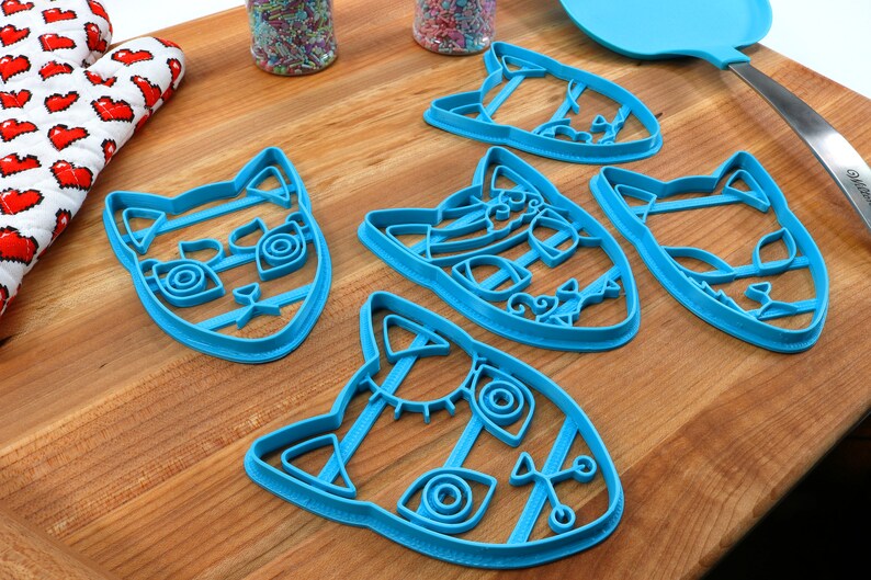 K&ouml;nnte beinhalten: Vier blaue, plastikene Ausstecher in Katzenform auf einer Holzoberfl&auml;che. Jeder Ausstecher hat ein anderes Design mit filigranen Details.
