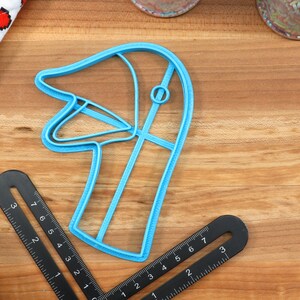 May include: A blue plastic cookie cutter shaped like a goose head. The cutter is on a wooden surface.