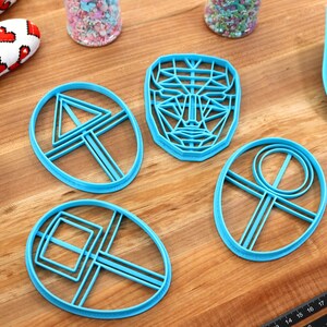 May include: Four blue plastic cookie cutters on a wooden surface. The cutters are shaped like a face, a triangle, a square, and a circle.