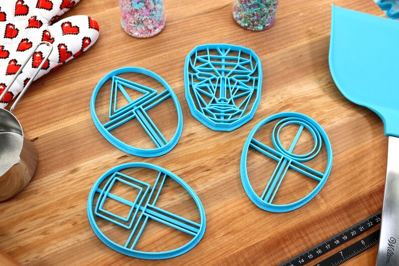 May include: Four blue plastic cookie cutters on a wooden surface. The cutters are shaped like a face, a triangle, a square, and a circle.