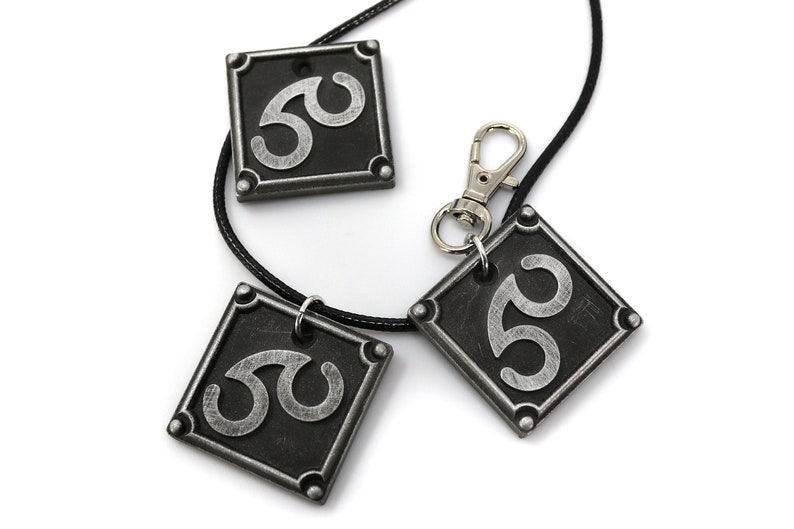 May include: Three silver-colored pendants with a black background and a stylized symbol. The pendants are attached to a black cord and a silver clasp.