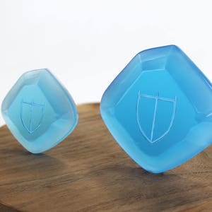 May include: Two blue glass paperweights with a shield design. The larger paperweight is on a wooden surface.