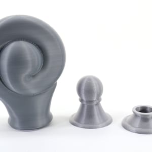 K&ouml;nnte beinhalten: Drei graue 3D-gedruckte Objekte werden vor einem wei&szlig;en Hintergrund gezeigt. Die Objekte umfassen ein gro&szlig;es, gewirbeltes Design, eine kleine abgerundete Oberseite und eine Basis mit einer kreisf&ouml;rmigen &Ouml;ffnung. Die Objekte sind wahrscheinlich dekorativ oder funktional.