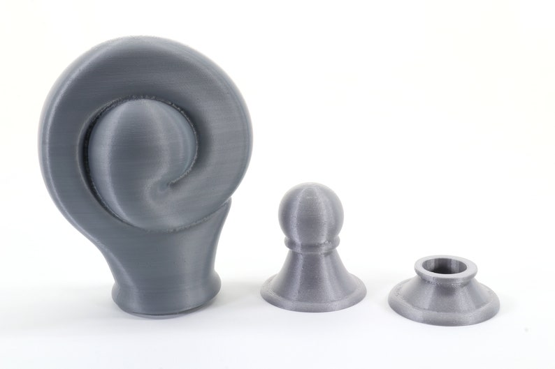K&ouml;nnte beinhalten: Drei graue 3D-gedruckte Objekte werden vor einem wei&szlig;en Hintergrund gezeigt. Die Objekte umfassen ein gro&szlig;es, gewirbeltes Design, eine kleine abgerundete Oberseite und eine Basis mit einer kreisf&ouml;rmigen &Ouml;ffnung. Die Objekte sind wahrscheinlich dekorativ oder funktional.
