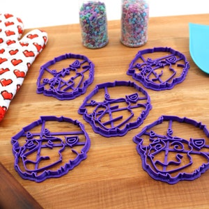 May include: Five purple plastic cookie cutters shaped like a dog's face. The cutters are on a wooden surface with a rolling pin, a red oven mitt, and two jars of sprinkles.