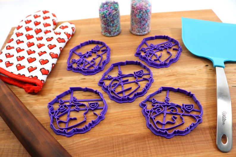 May include: Five purple plastic cookie cutters shaped like a dog's face. The cutters are on a wooden surface with a rolling pin, a red oven mitt, and two jars of sprinkles.