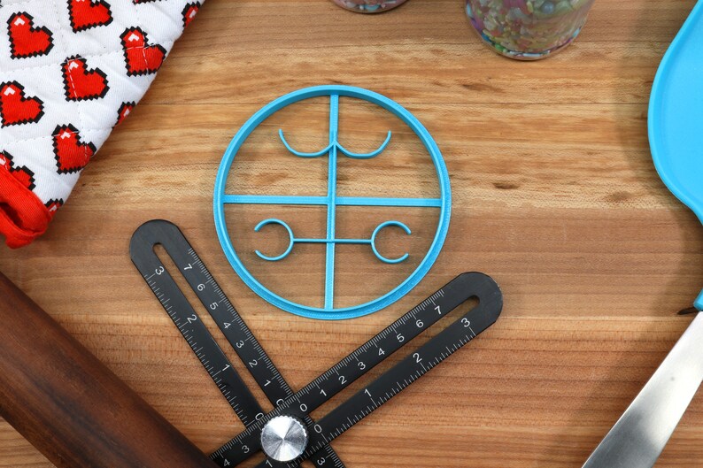 May include: A blue plastic cookie cutter shaped like a circle with four smaller circles inside. The cookie cutter is on a wooden surface.