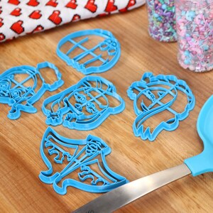May include: A set of six blue plastic cookie cutters shaped like cartoon characters. The cutters are on a wooden surface with a rolling pin and sprinkles in the background.