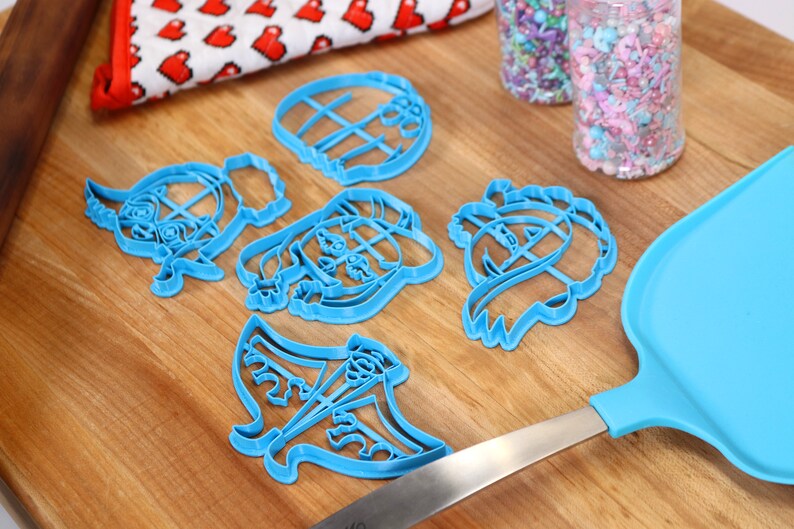 May include: A set of six blue plastic cookie cutters shaped like cartoon characters. The cutters are on a wooden surface with a rolling pin and sprinkles in the background.
