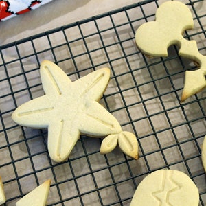 Op de afbeelding: Een close-up van een draadrek met zes koekjes in verschillende vormen, waaronder een ster, een hart, een sleutelgat en een cirkel. De koekjes zijn allemaal lichtbruin van kleur en hebben een licht glanzende afwerking.