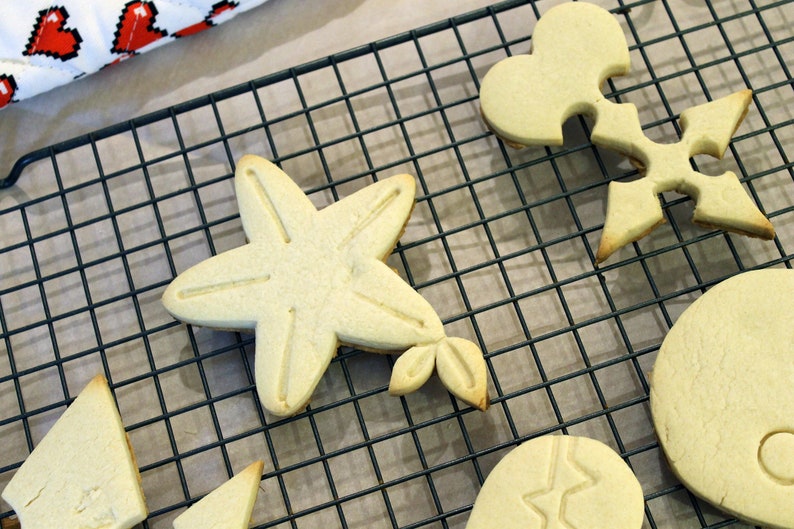 Op de afbeelding: Een close-up van een draadrek met zes koekjes in verschillende vormen, waaronder een ster, een hart, een sleutelgat en een cirkel. De koekjes zijn allemaal lichtbruin van kleur en hebben een licht glanzende afwerking.