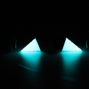 May include: Two glowing, triangular objects emit a bright turquoise light against a black background. The objects appear to be illuminated from within, casting a soft glow and creating a dramatic effect. The objects are likely decorative or artistic.