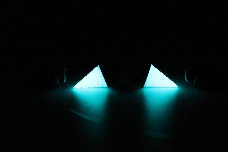 May include: Two glowing, triangular objects emit a bright turquoise light against a black background. The objects appear to be illuminated from within, casting a soft glow and creating a dramatic effect. The objects are likely decorative or artistic.