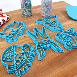 May include: A collection of blue plastic cookie cutters in various shapes, including a heart, a star, and a character with a hat. A red and white oven mitt with heart patterns is on the left. Sprinkles and a blue spatula are also present.