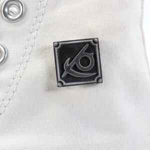May include: White canvas high-top sneaker with a silver metal eyelet detail and a black and silver metal pin with a stylized eye and blade design.
