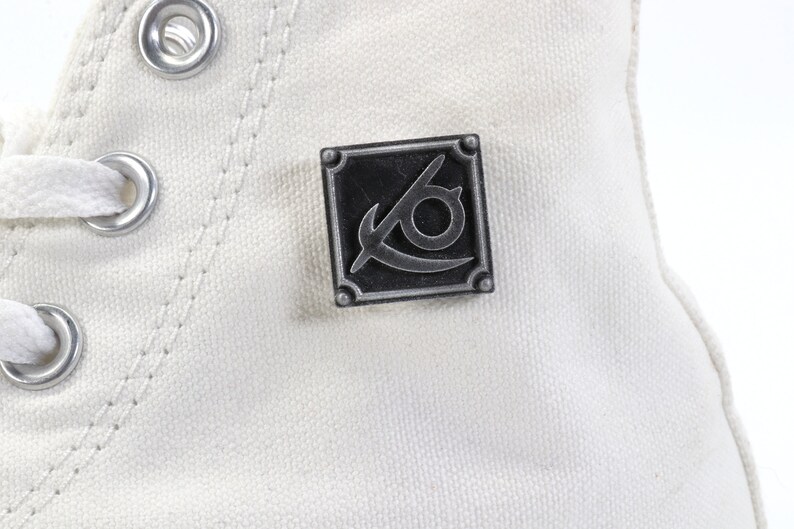 May include: White canvas high-top sneaker with a silver metal eyelet detail and a black and silver metal pin with a stylized eye and blade design.