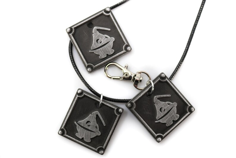 May include: Three silver-toned pendants with a black and white image of a character wearing a hat. The pendants are attached to a black cord and a silver-toned clasp.
