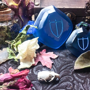 May include: Two blue glass dice with a shield symbol etched on each face. The dice are on a dark patterned surface with dried flowers and other natural objects.