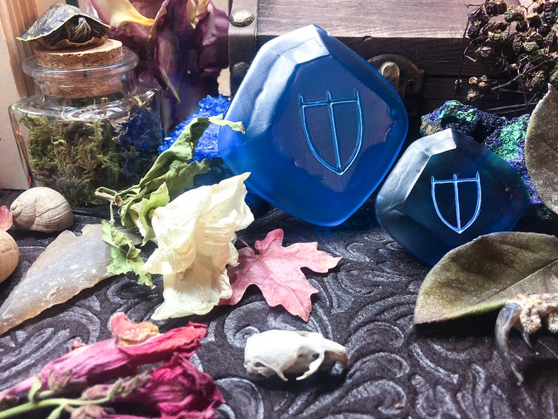 May include: Two blue glass dice with a shield symbol etched on each face. The dice are on a dark patterned surface with dried flowers and other natural objects.