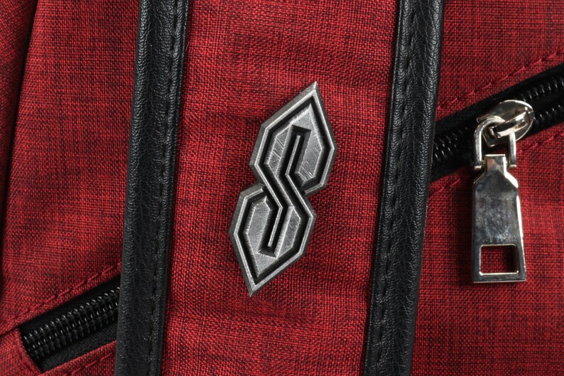 May include: Close-up of a red backpack with a textured fabric. A silver-colored emblem in the shape of a stylized "S" is affixed to the front. Black leather trim and a silver zipper are also visible.
