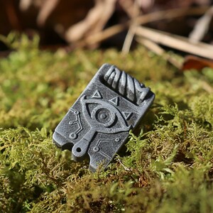 May include: A silver-colored metal pendant with a detailed design of an eye and triangles. The pendant is on a bed of green moss.