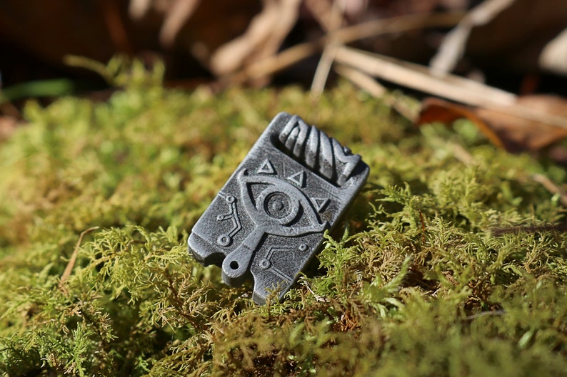 May include: A silver-colored metal pendant with a detailed design of an eye and triangles. The pendant is on a bed of green moss.