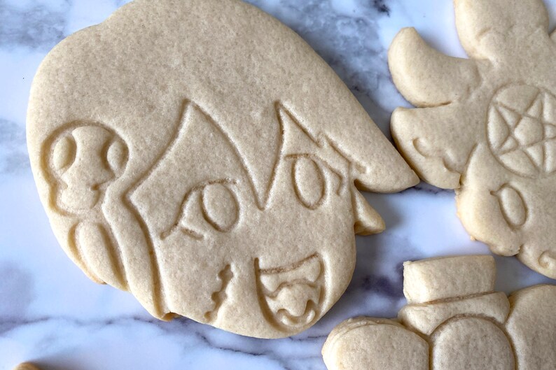 Pode incluir: Um close-up de um biscoito de a&ccedil;&uacute;car decorado com um personagem de estilo anime com olhos grandes e um sorriso largo. O biscoito &eacute; de cor marrom claro e tem uma superf&iacute;cie ligeiramente texturizada.