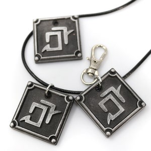 K&ouml;nnte beinhalten: Drei quadratische Anh&auml;nger in Silber mit einem schwarzen Symbol auf jedem. Die Anh&auml;nger sind an einer schwarzen Schnurkette mit einem silbernen Verschluss befestigt.