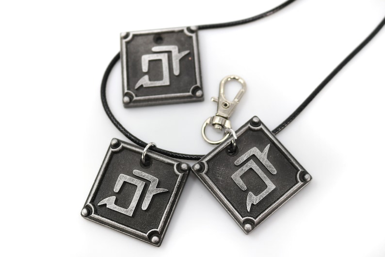 K&ouml;nnte beinhalten: Drei quadratische Anh&auml;nger in Silber mit einem schwarzen Symbol auf jedem. Die Anh&auml;nger sind an einer schwarzen Schnurkette mit einem silbernen Verschluss befestigt.