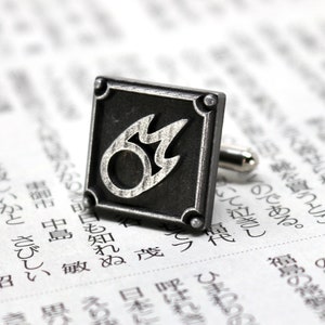 K&ouml;nnte beinhalten: Silberne Manschettenkn&ouml;pfe mit einem schwarzen quadratischen Design, das eine stilisierte silberne Flamme zeigt.