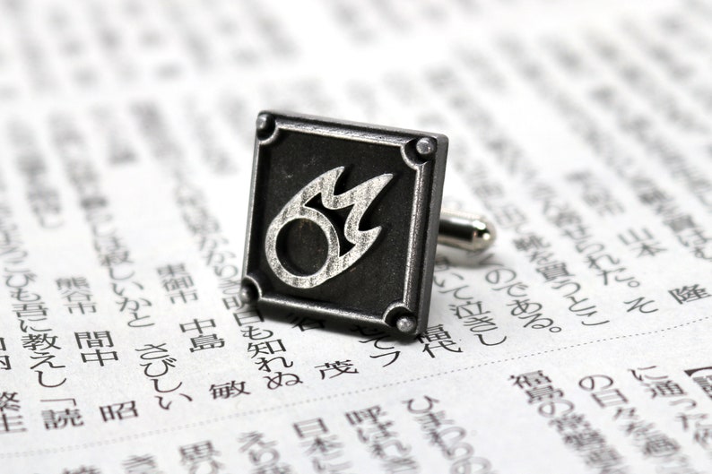 K&ouml;nnte beinhalten: Silberne Manschettenkn&ouml;pfe mit einem schwarzen quadratischen Design, das eine stilisierte silberne Flamme zeigt.