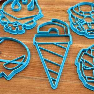 May include: Five blue plastic cookie cutters on a wooden surface. The cutters are shaped like a ghost, an ice cream cone, a donut, a skull, and a robot.