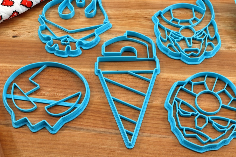 May include: Five blue plastic cookie cutters on a wooden surface. The cutters are shaped like a ghost, an ice cream cone, a donut, a skull, and a robot.