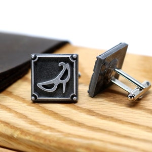May include: A pair of silver cufflinks with a square design. One cufflink features a stylized image of a high heel shoe. The other cufflink is plain.
