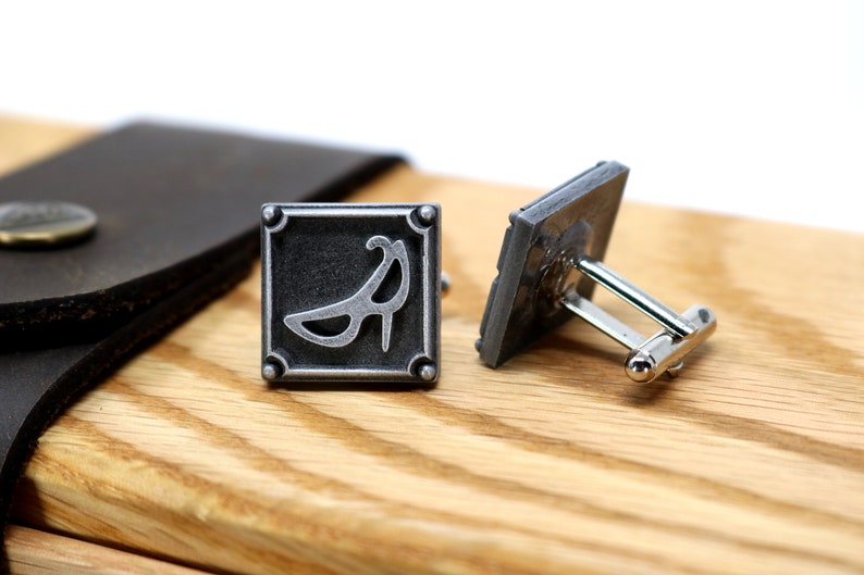 May include: A pair of silver cufflinks with a square design. One cufflink features a stylized image of a high heel shoe. The other cufflink is plain.
