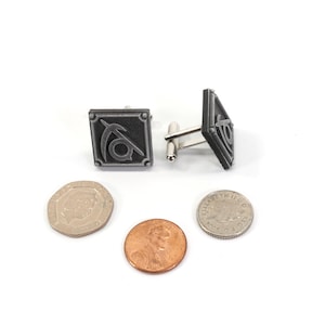 May include: A pair of silver cufflinks with a black and silver design. The design features a stylized symbol with a curved line and a circle. The cufflinks are on a white surface with three coins.