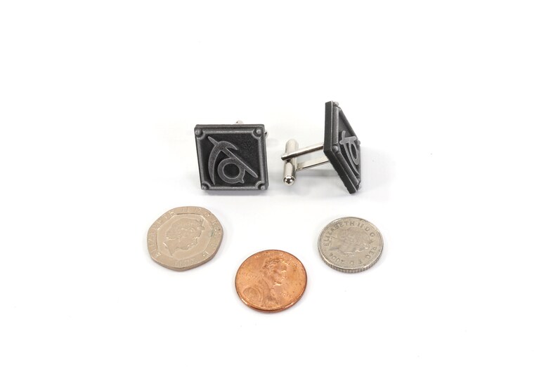 May include: A pair of silver cufflinks with a black and silver design. The design features a stylized symbol with a curved line and a circle. The cufflinks are on a white surface with three coins.