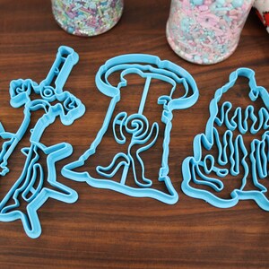 Puede incluir: Tres cortadores de galletas de pl&aacute;stico azul con forma de &aacute;rbol, casa y unicornio. Los cortadores est&aacute;n sobre una superficie de madera.